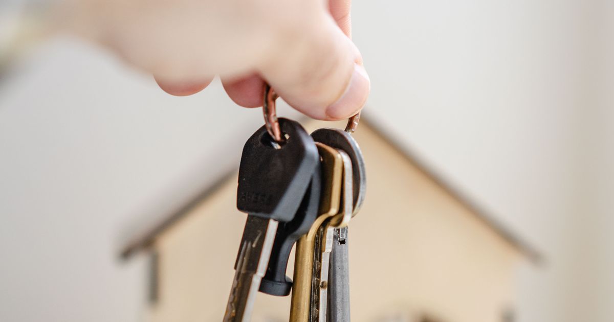 A hand holding house keys in front of a small wooden model house