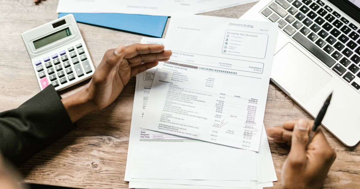 Hands holding a printed bank statement next to a calculator and a laptop